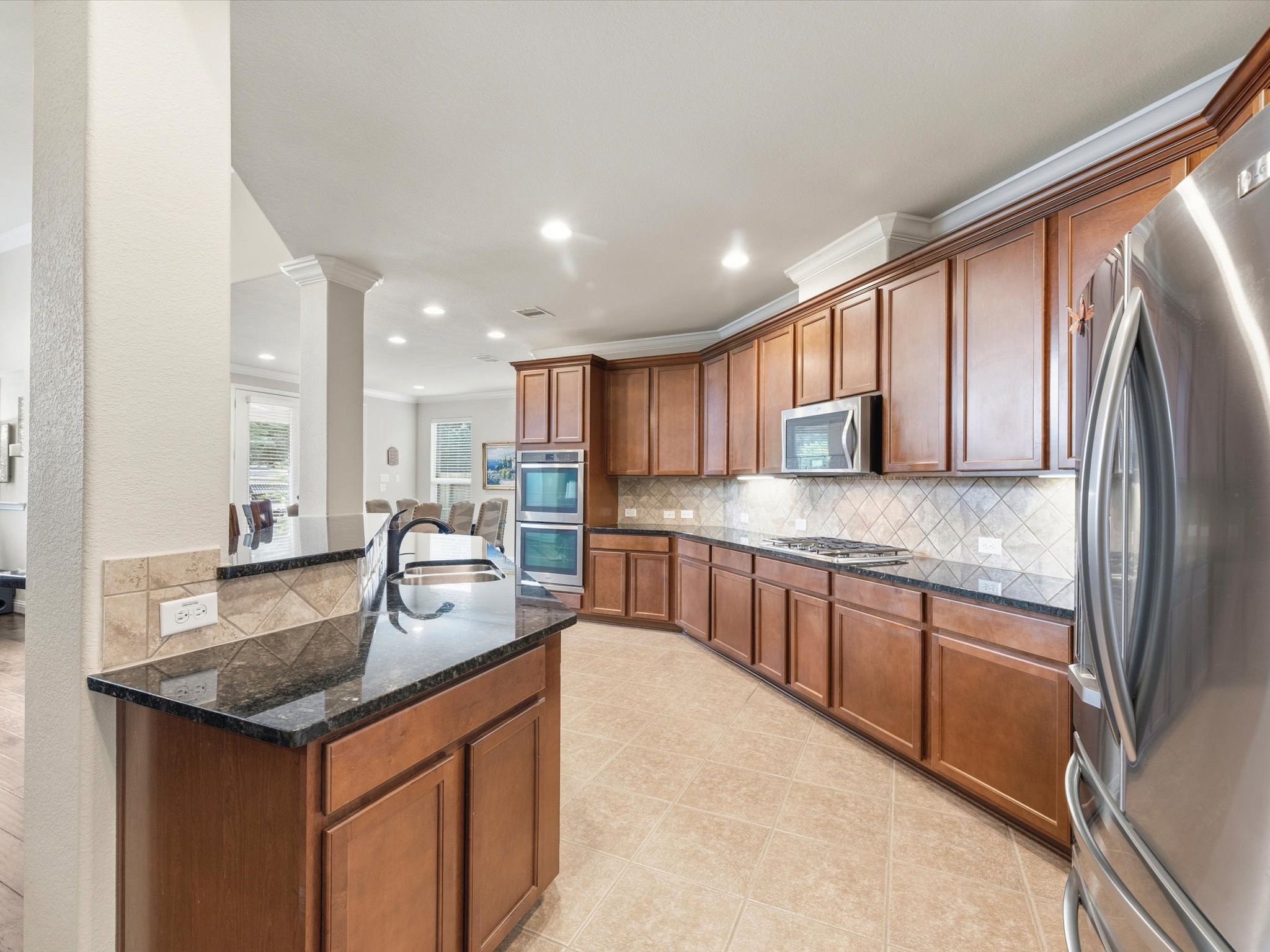 3907 Randle Ridge Court Fulshear, TX 77441 - Photo 13 of 46 This photo showcases a spacious kitchen featuring dark granite countertops, ample wooden cabinetry, stainless steel appliances, and a tiled backsplash. The open layout connects to a dining area, creating a welcoming space for entertaining.