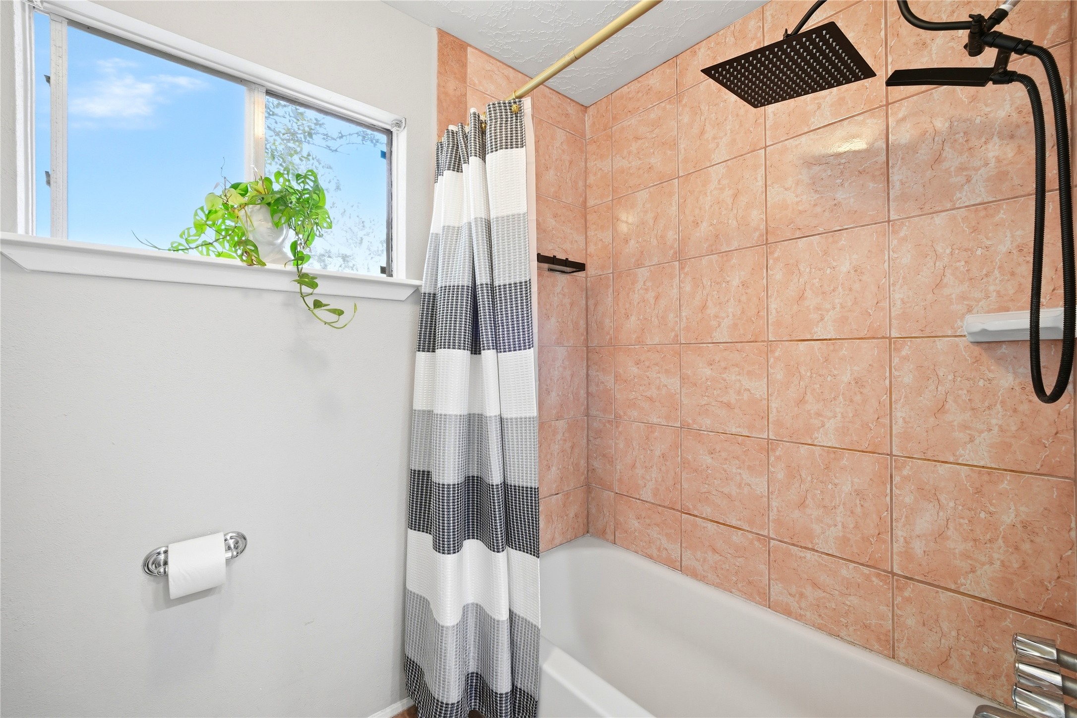 4718 Temple Bell Drive Spring, TX 77388 - Photo 36 of 50 Cozy bathroom with peach-toned tiles, a modern rain showerhead, and a white bathtub. A striped shower curtain adds style, while a small window with a plant offers natural light.
