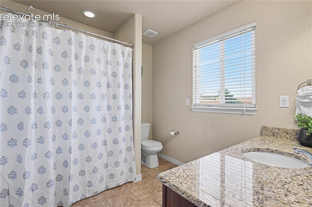 6598 Alliance Loop Colorado Springs, CO 80925 - Photo 23 of 39 Bathroom featuring a shower area, a single vanity with a granite-style countertop and integrated sink, a built-in toilet, tiled flooring, and a window with blinds