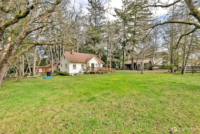 a view of a white house in front of a big yard with plants and large trees
