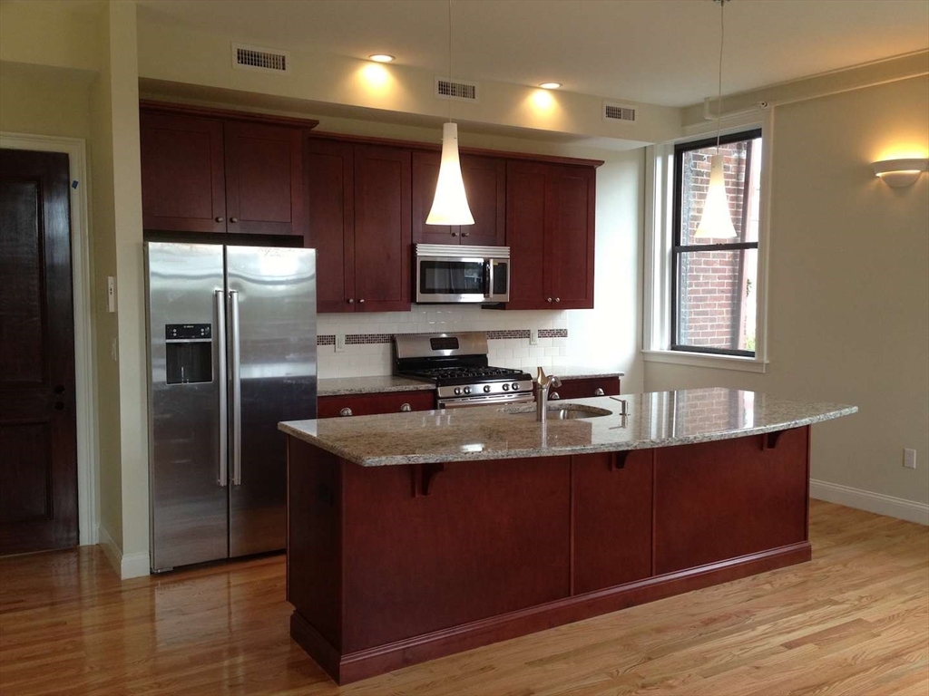 191 Main Street, Unit 2B Gloucester, MA 01930 - Photo 6 of 20 a kitchen with kitchen island granite countertop wooden cabinets a refrigerator and a sink