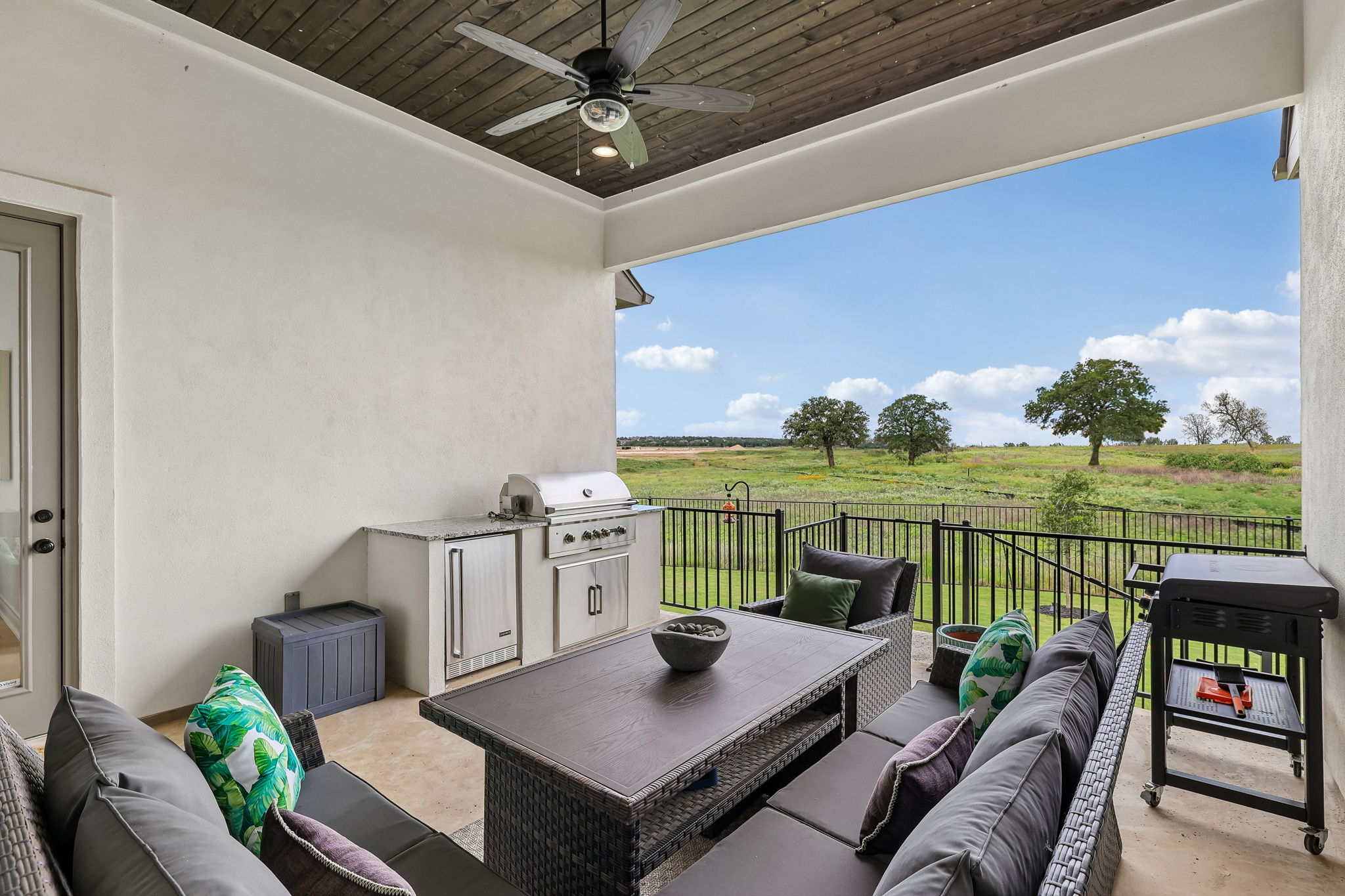 130 Firethorn Loop Bastrop, TX 78602 - Photo 4 of 38 Covered outdoor patio featuring a wood-finish ceiling with a ceiling fan, an outdoor kitchen with a built-in grill and refrigerator, a concrete patio surface, and a black metal fence enclosing the space