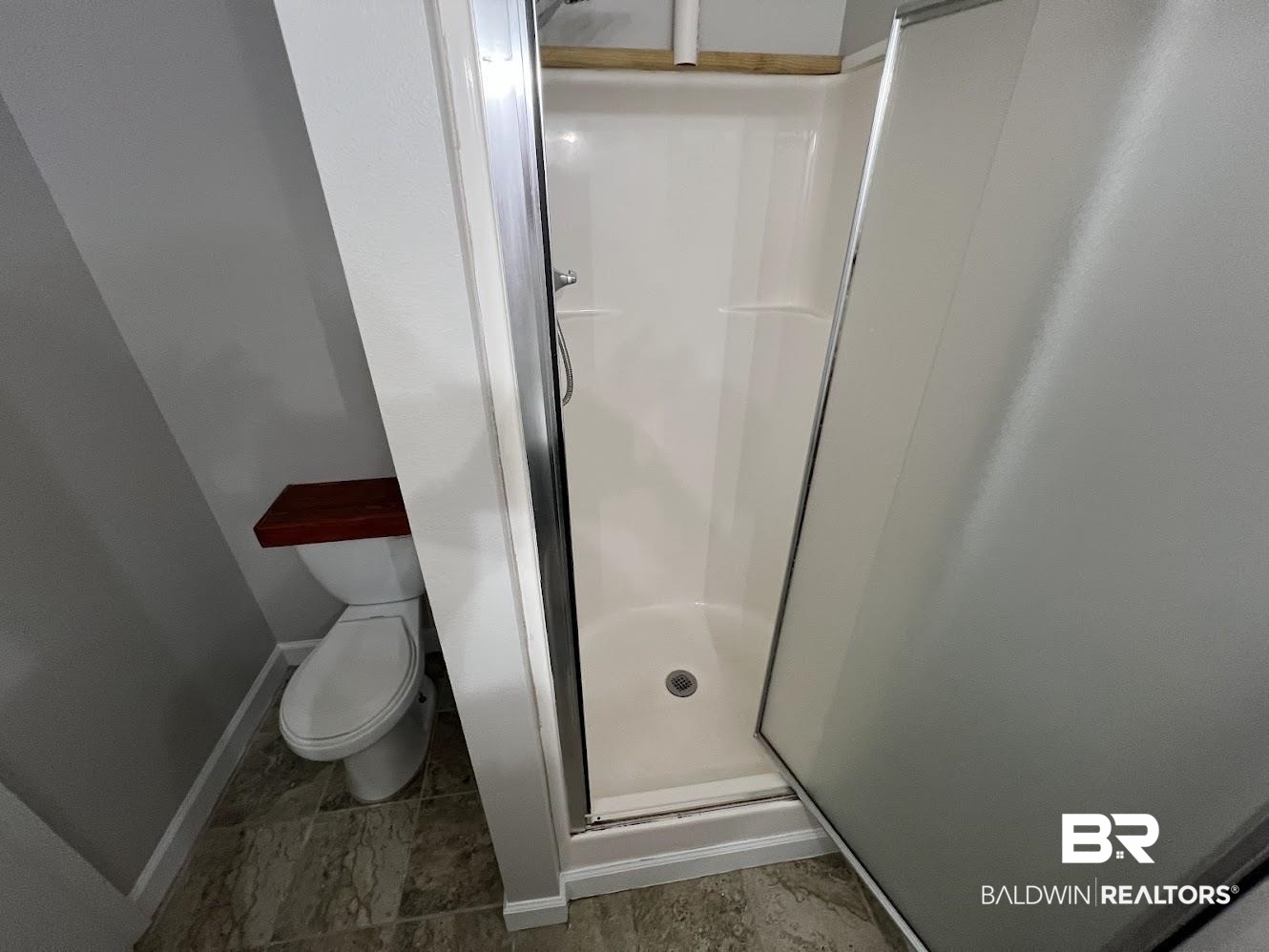 266 Ridgewood Drive Daphne, AL 36526 - Photo 24 of 34 Compact bathroom featuring a stand-up shower with frosted glass doors, white toilet with a wood-finish tank lid, light gray walls, and patterned tile flooring