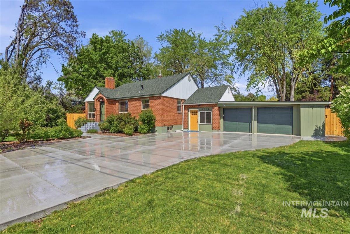 Brick home with a green shingle roof, featuring a multi-panel concrete driveway, a two-car garage with green doors, an additional covered parking space, and mature perimeter landscaping