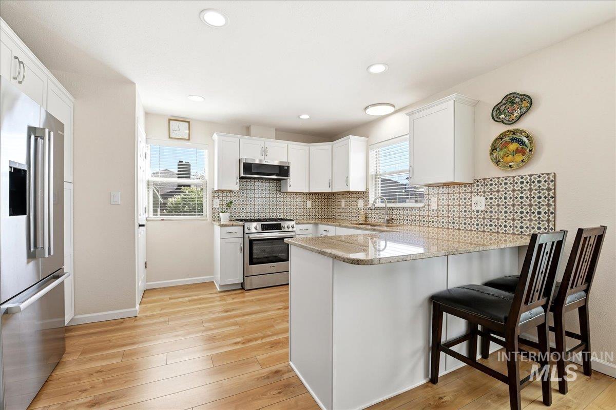 6710 West Ustick Road Boise, ID 83704 - Photo 13 of 42 Bright kitchen featuring white cabinetry, stainless steel appliances, patterned tile backsplash, wood-finish flooring, and a peninsula with countertop seating