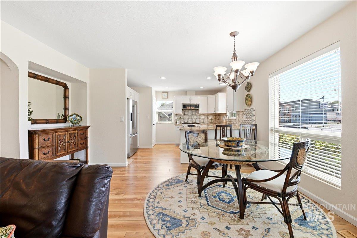 6710 West Ustick Road Boise, ID 83704 - Photo 14 of 42 Open-concept dining area featuring wood-finish flooring, a contemporary chandelier, a large window with blinds, and a view into the kitchen with white cabinetry and stainless steel appliances