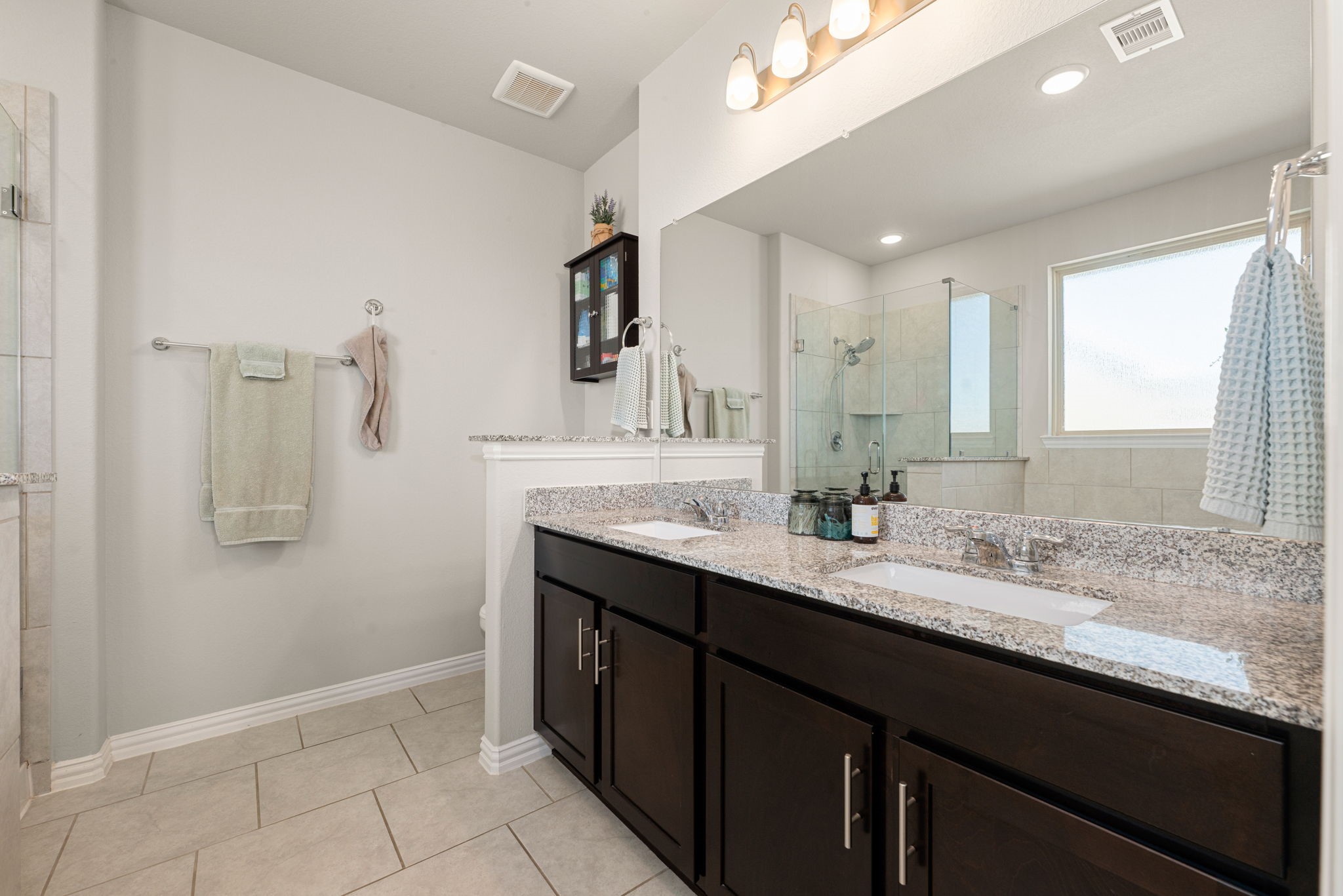 8414 Divot Trace Fulshear, TX 77441 - Photo 14 of 25 This spa-inspired primary bathroom boasts a dual-sink vanity with rich espresso cabinetry and sleek white countertops. A large, wall-to-wall mirror reflects the bright recessed lighting and modern fixtures, enhancing the room's airy feel. The clean lines are complemented by neutral tile flooring and a dedicated space for a walk-in shower or soaking tub area.
