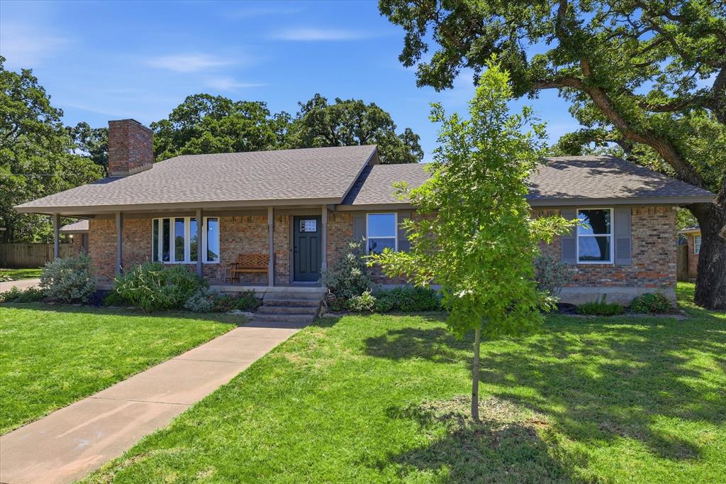 Brick exterior featuring a covered front porch, grey shingle roof, and mature trees providing shade