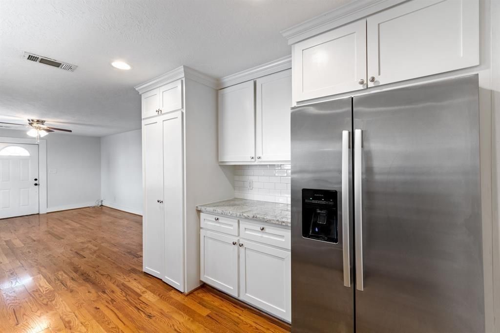 1305 Idylwild Street Houston, TX 77009 - Photo 12 of 31 This photo showcases a bright kitchen with white cabinetry, granite countertops, and a stainless steel refrigerator. It opens up to a living area with hardwood floors and a ceiling fan, creating a welcoming and modern space.