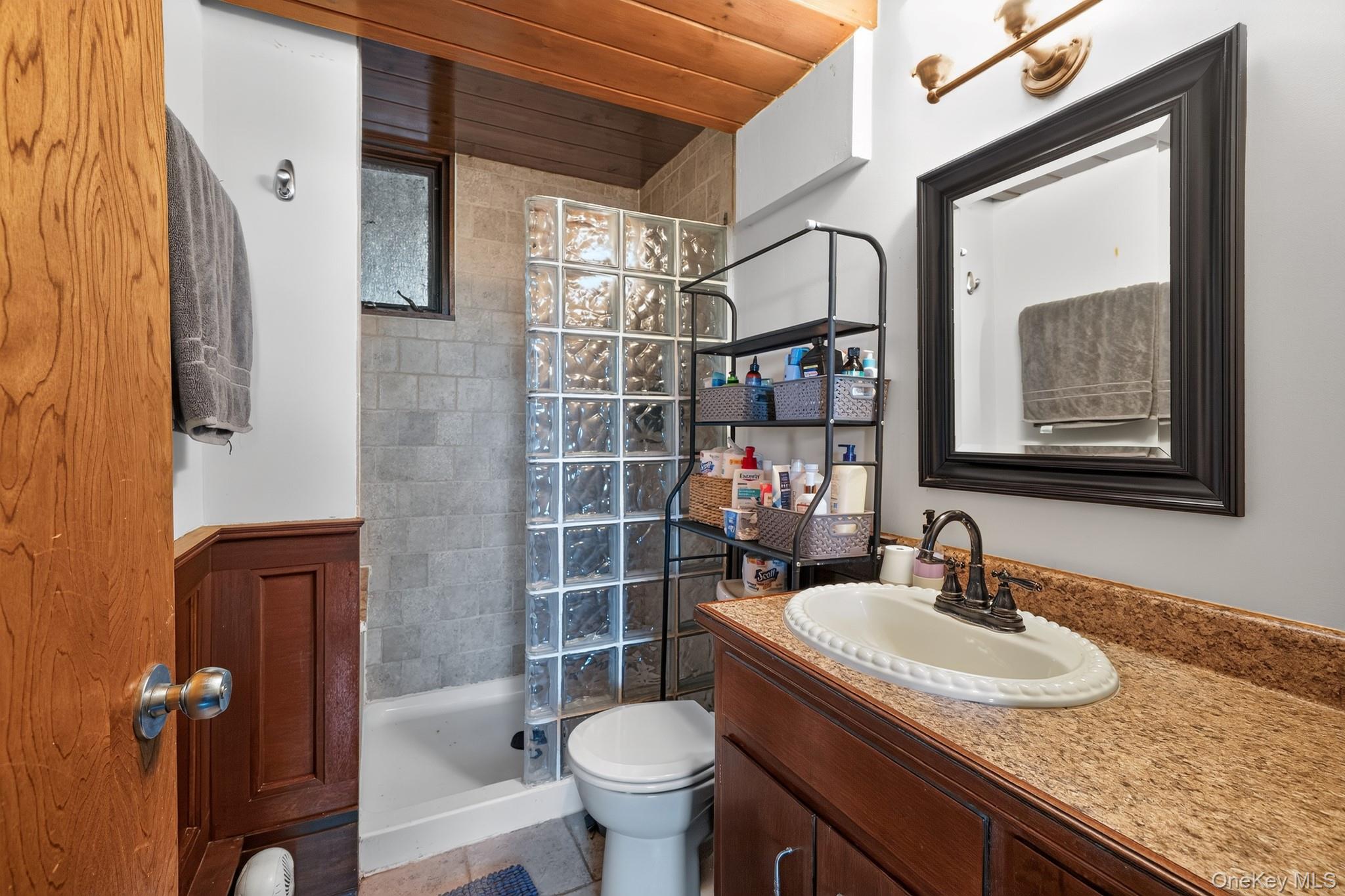 424 Laroe Road Chester, NY 10918 - Photo 7 of 43 Bathroom featuring a wood-panel ceiling, glass block shower enclosure, tiled shower surround, wood-finish vanity with a built-in sink, and a framed mirror