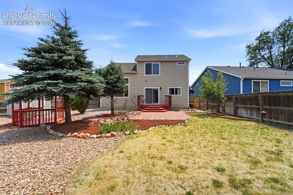 7904 Coffee Road Peyton, CO 80831 - Photo 36 of 48 Rear exterior featuring a brick paver patio, wood deck with red railing, and a gravel-covered area with mature trees