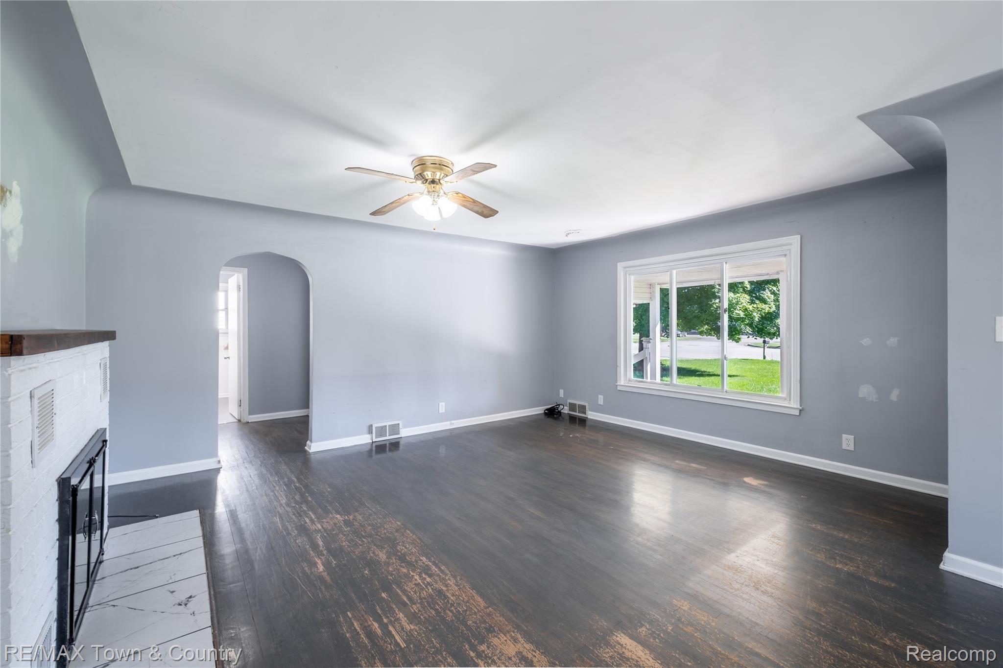 300 Elms Road Flushing, MI 48433 - Photo 7 of 26 Hardwood flooring, white brick fireplace with dark wood mantel, arched entryway, ceiling fan with light fixture, and large window with white trim