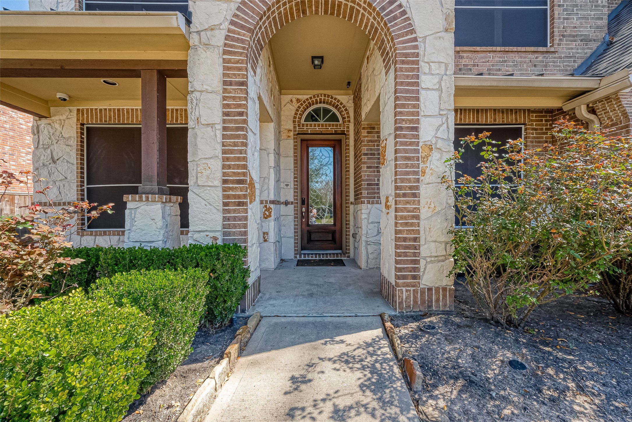 Charming front entry with a stone and brick facade, featuring an arched doorway and well-maintained shrubs, creating an inviting entrance.