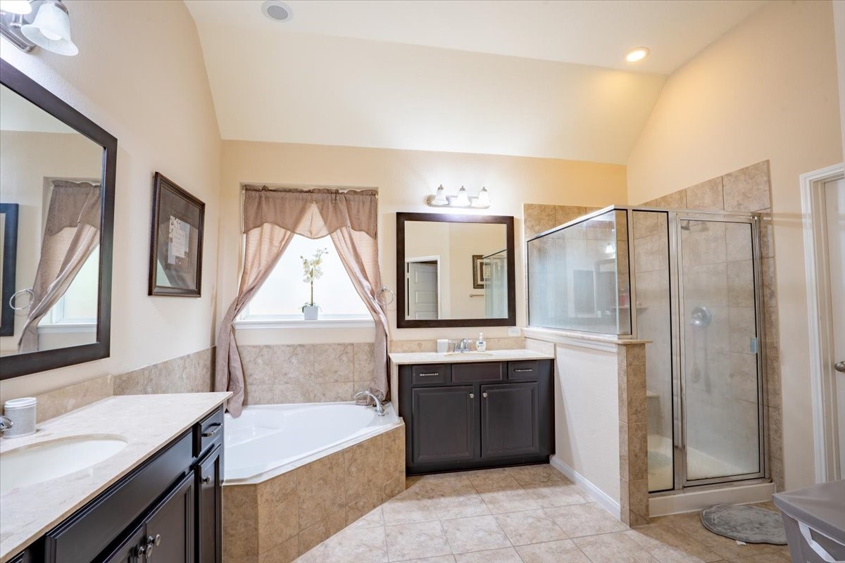 22002 Flashing Ridge Spring, TX 77389 - Photo 17 of 43 This bathroom features a spacious layout with a large soaking tub, glass-enclosed shower, dual sinks with dark cabinetry, and neutral tile flooring. Soft lighting and a window with curtains add a warm, inviting touch.
