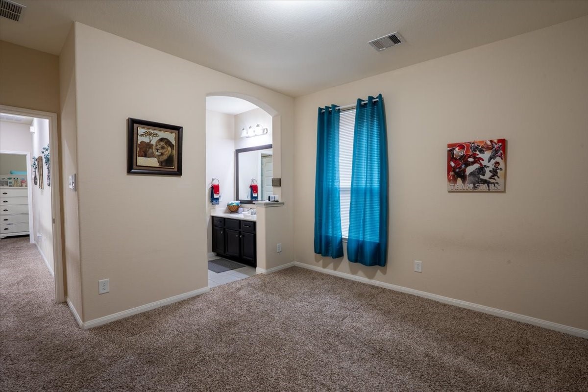22002 Flashing Ridge Spring, TX 77389 - Photo 31 of 43 This photo shows a cozy bedroom with carpeted flooring, neutral walls, and a window with blue curtains. An open doorway leads to a bathroom with a dark vanity and ample lighting. A hallway connects to another room with visible furniture.