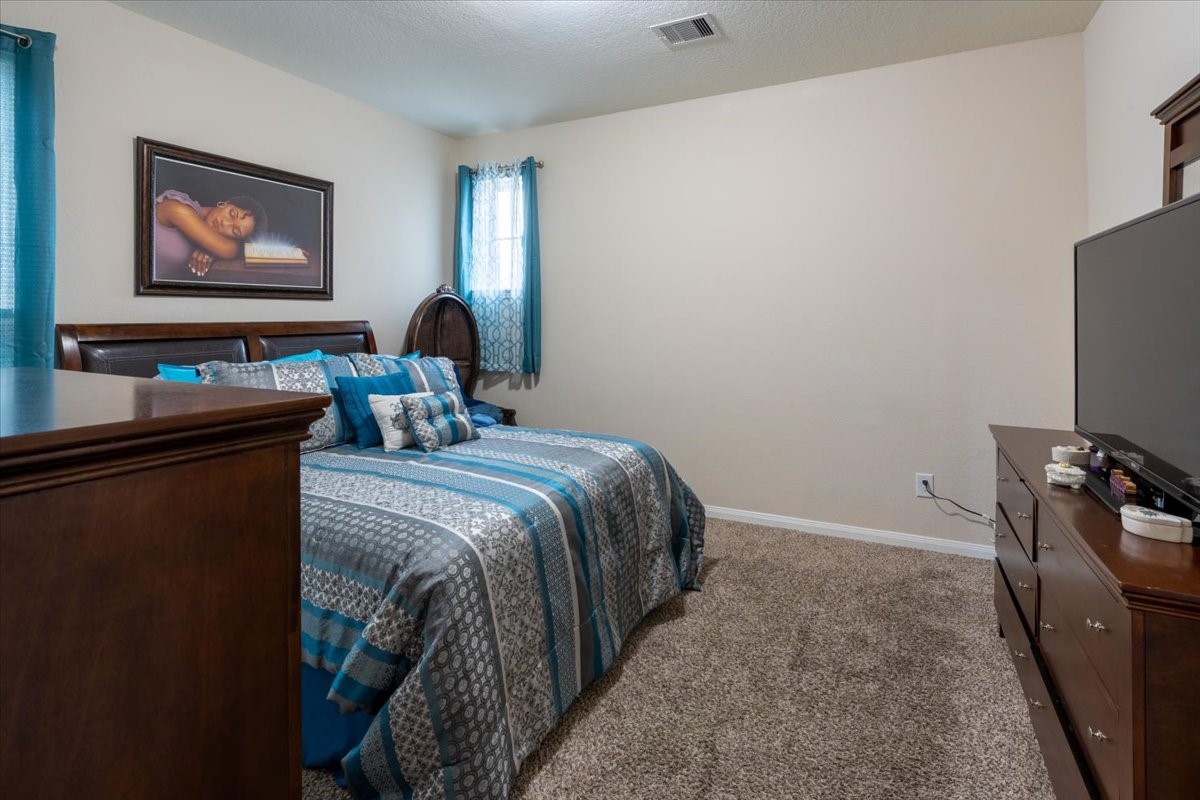 22002 Flashing Ridge Spring, TX 77389 - Photo 33 of 43 This bedroom features a cozy setup with a large bed adorned in blue and gray bedding. The room includes plush carpet, a wooden dresser with a TV, and a window with teal curtains, offering a comfortable and inviting atmosphere.