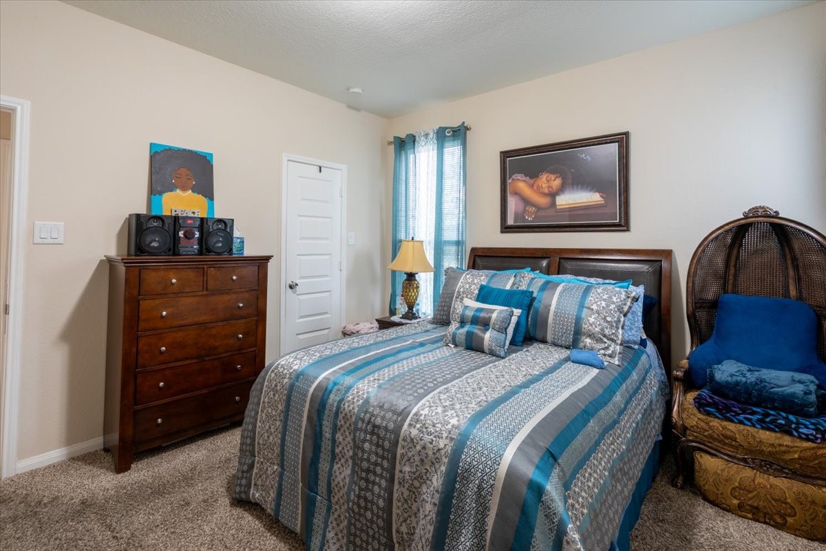 22002 Flashing Ridge Spring, TX 77389 - Photo 35 of 43 Cozy bedroom with a double bed featuring blue and gray bedding, a wooden dresser, and a wicker chair. The room is accented with artwork and a soft carpet, creating a warm and inviting atmosphere.