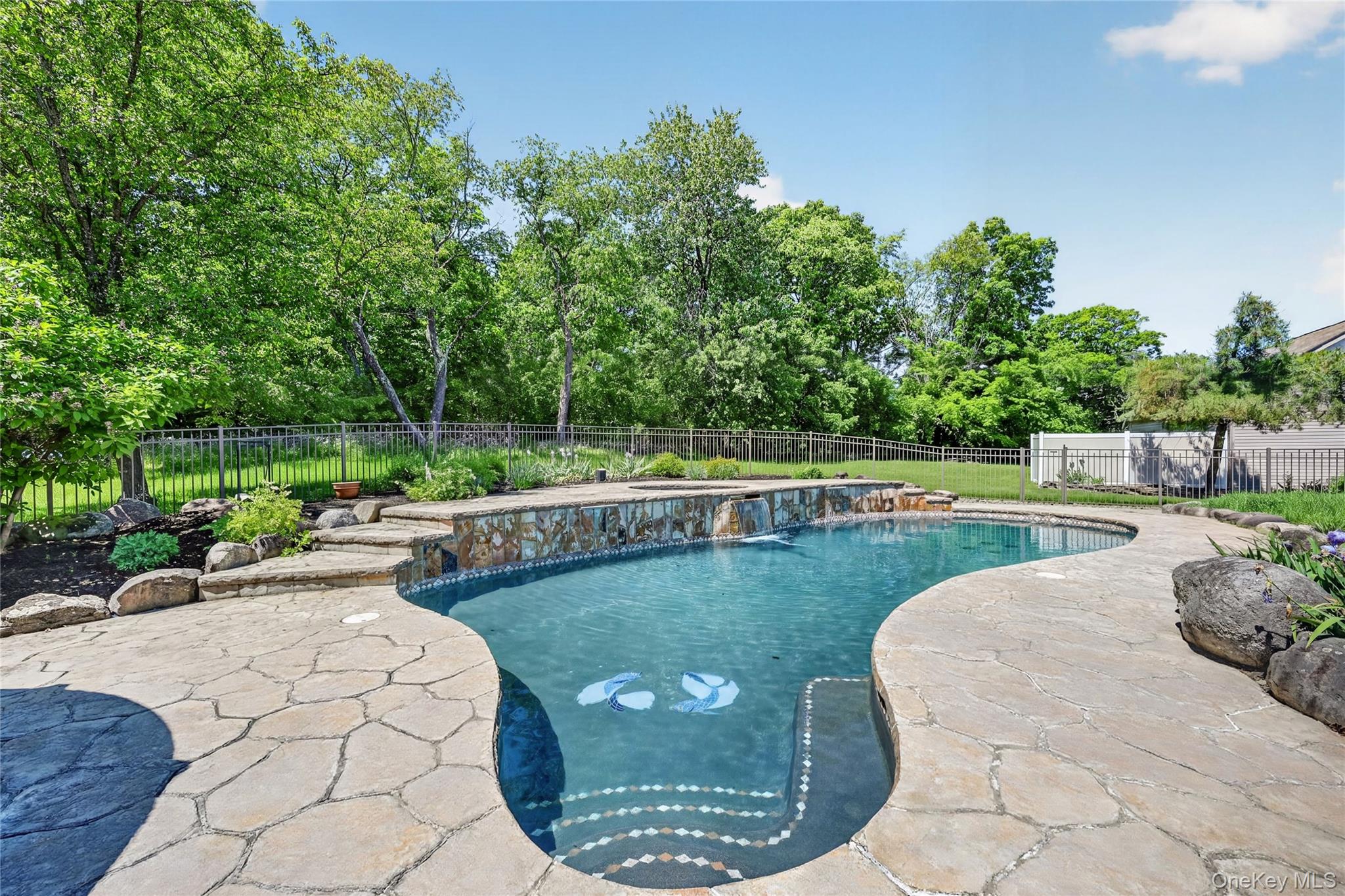 954 Ridgebury Road New Hampton, NY 10958 - Photo 38 of 45 Surrounded by mature greenery and gentle water features, the pool area feels peaceful, private, and resort-like. The heated gunite pool is the centerpiece of this backyard retreat, complete with a raised hot tub and three beautiful waterfalls that cascade over the wall into the pool below. It is an inviting space designed for summer days, evening swims, and unforgettable gatherings at home.