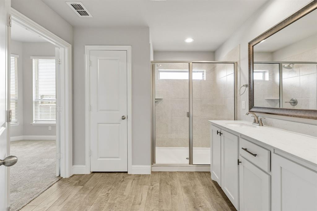 915 Timber Ridge Drive Justin, TX 76247 - Photo 23 of 39 Bathroom featuring a spacious shower with glass enclosure, dual white vanity with undermount sink, stone countertop, wood-finish flooring, and a large framed mirror