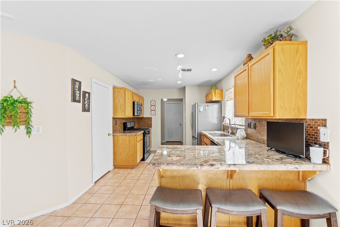 1419 Red Sunset Avenue Henderson, NV 89074 - Photo 12 of 35 Kitchen with a peninsula, stainless steel appliances, light tile patterned floors, backsplash, and light wood finish cabinetry