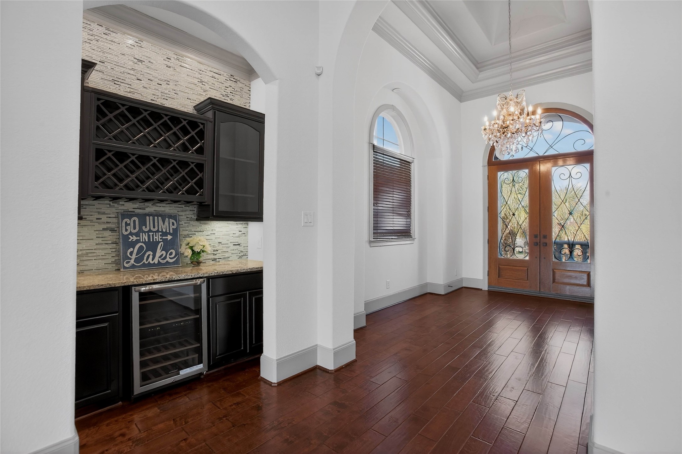 12349 Tramonto Conroe, TX 77304 - Photo 18 of 50 An elegant entryway with a chandelier, arched wooden double doors, and hardwood floors. To the left, there's a stylish wet bar with wine storage and a mini-fridge, featuring dark cabinetry and a stone backsplash.