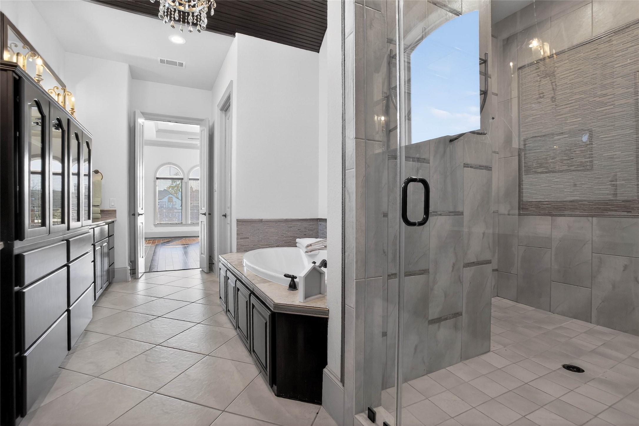 12349 Tramonto Conroe, TX 77304 - Photo 23 of 50 This spacious bathroom features a large glass-enclosed shower, a luxurious soaking tub, and elegant dark wood cabinetry. The room is brightened by a chandelier and natural light streaming through the primary room with arched windows.