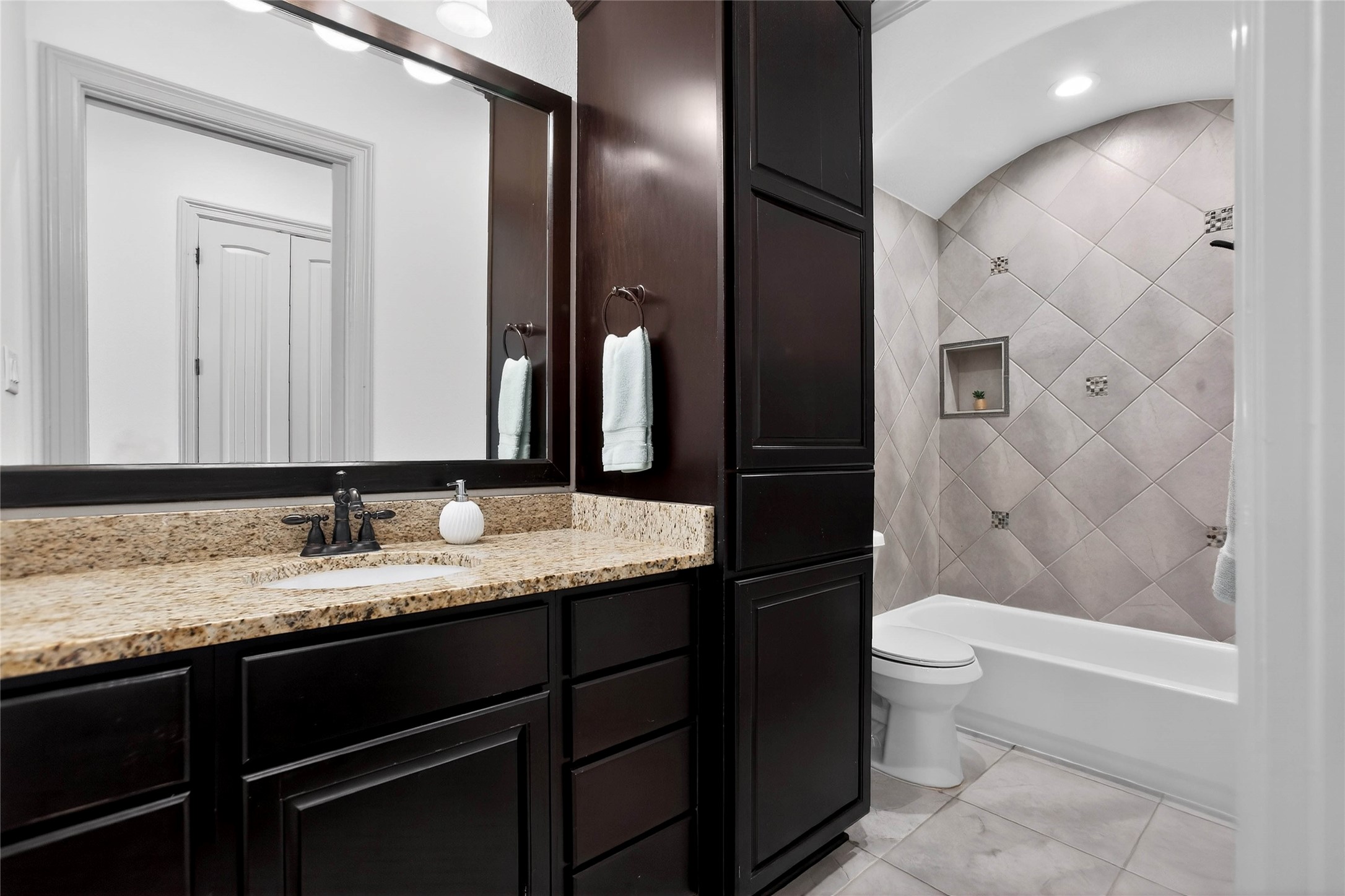 12349 Tramonto Conroe, TX 77304 - Photo 34 of 50 This bathroom features elegant dark wood cabinetry, a granite countertop, and a large mirror. The shower area has stylish tilework with built-in shelving, and the space is well-lit, creating a modern and inviting atmosphere.
