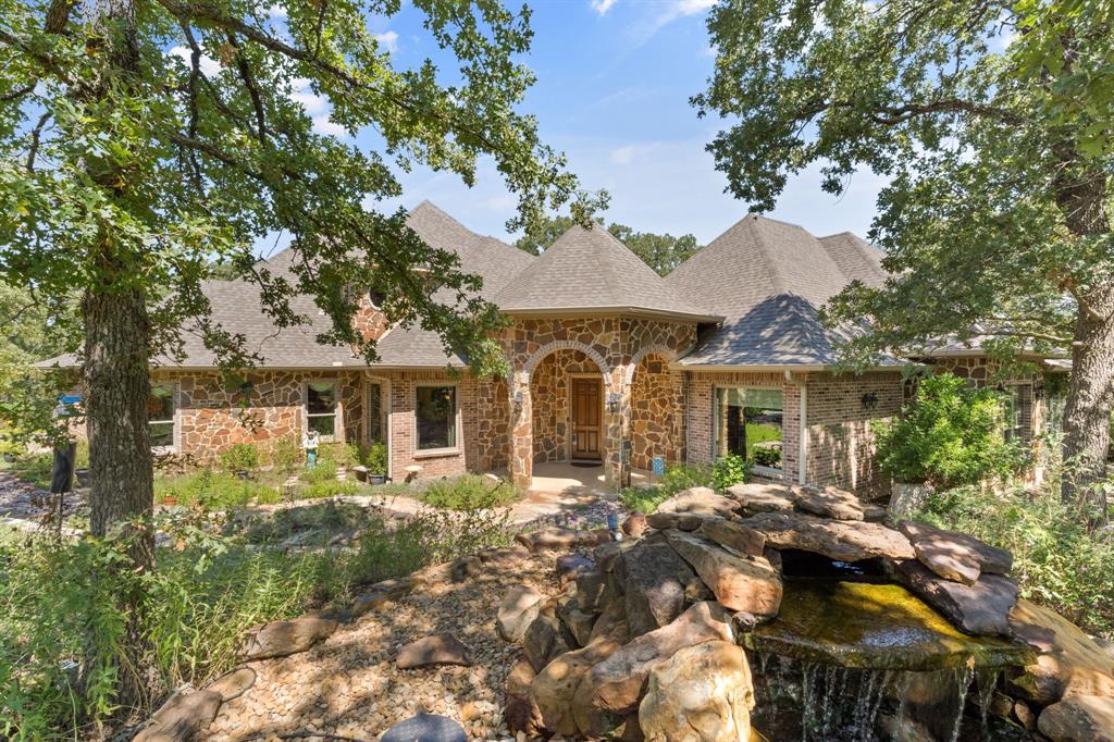View of front facade featuring a shingled roof, stone siding, a small pond, and brick siding