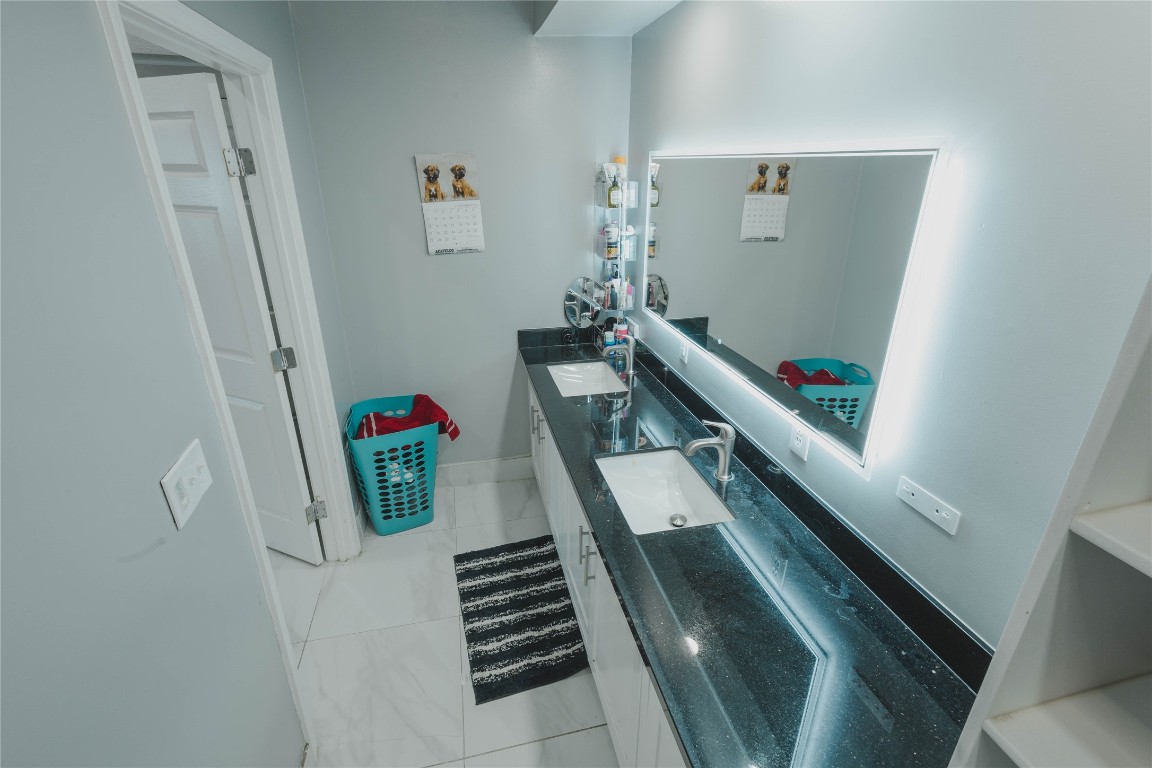 918 Fresa Road Pasadena, TX 77502 - Photo 17 of 47 Modern Hall bathroom with sleek black countertops, dual sinks, and a large, illuminated mirror. The space is bright with white tiled floors and light gray walls, offering a clean and contemporary feel.