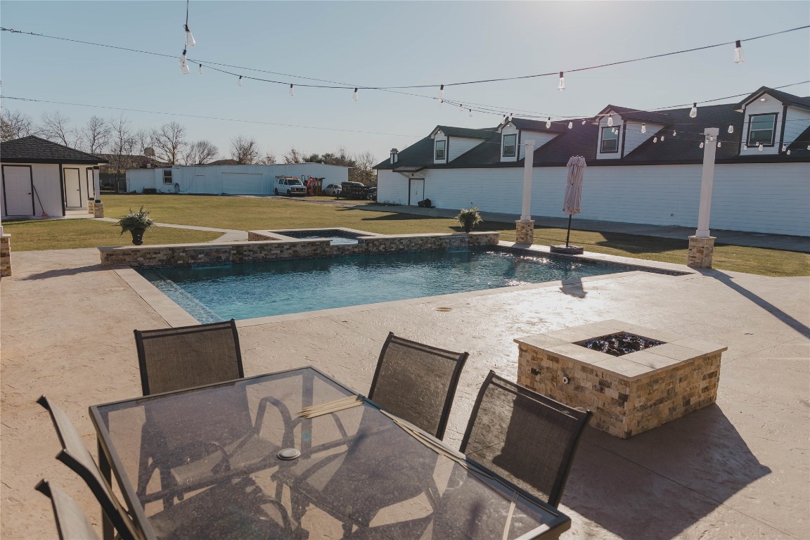 918 Fresa Road Pasadena, TX 77502 - Photo 22 of 47 This spacious backyard featuring a sleek pool and jacuzzi with a stone border, surrounded by a large patio area. There are a stylish outdoor dining set and a fire pit, perfect for entertaining. The setup is enhanced by string lights overhead, creating a cozy ambiance.