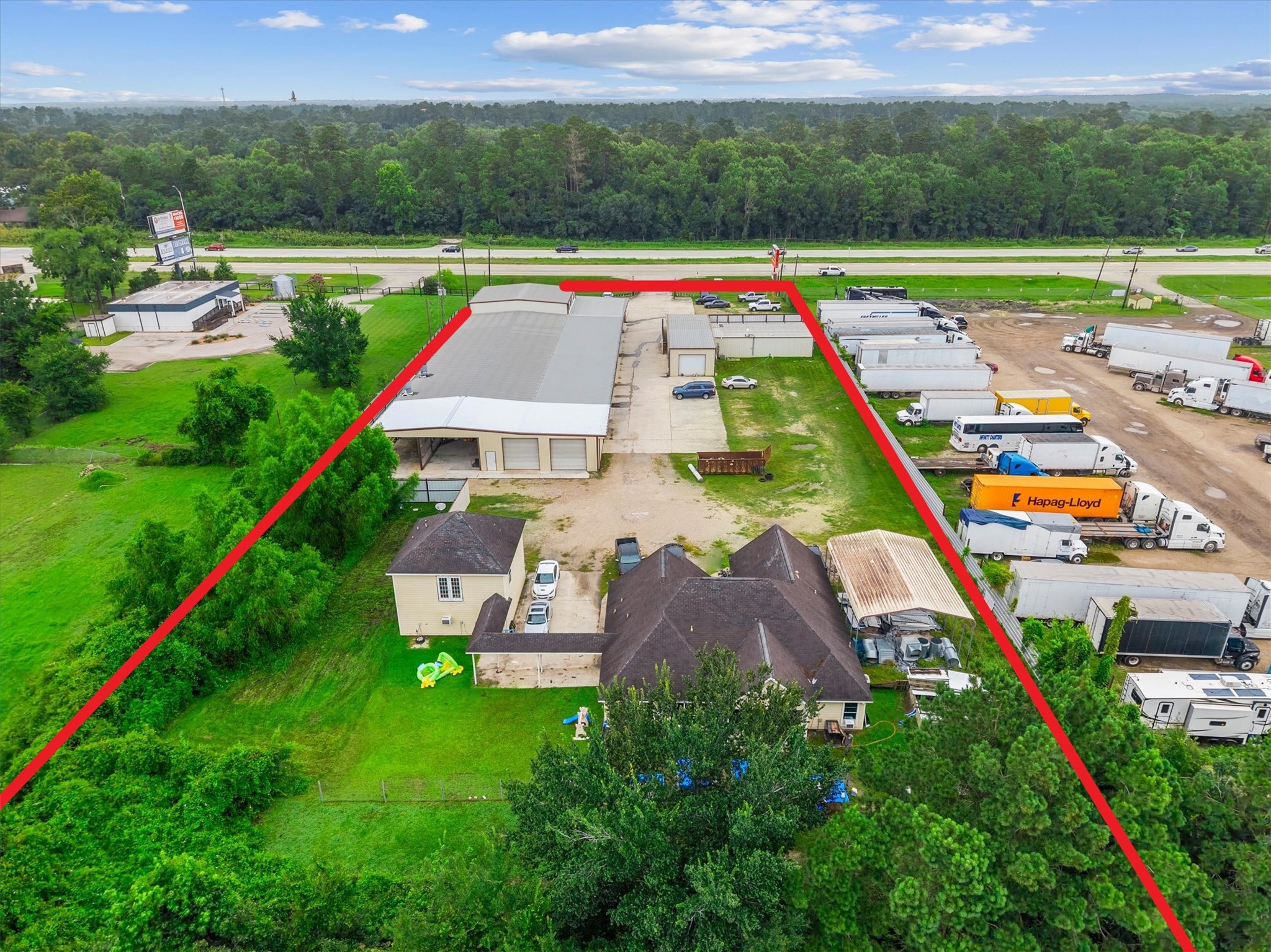 11066 Sh 242 Conroe, TX 77385 - Photo 6 of 21 This aerial photo shows a property with a residential home and a large industrial building, bordered by red lines. The area includes open green space and is adjacent to a highway, with trucks parked nearby, suggesting a potential mix of residential and commercial use.