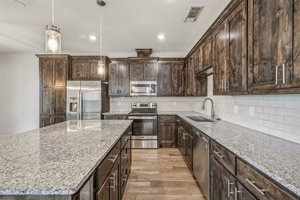 745 North Miller Avenue Stephenville, TX 76401 - Photo 11 of 31 Kitchen featuring a large central island with light-toned countertops, rich wood-finish cabinetry, stainless steel appliances, a white subway tile backsplash, and wood-finish flooring