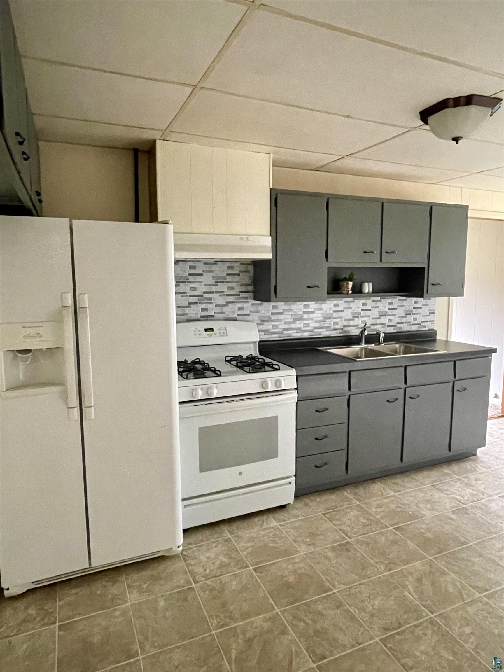 Kitchen with gray cabinetry, white appliances, backsplash, sink, and light tile floors