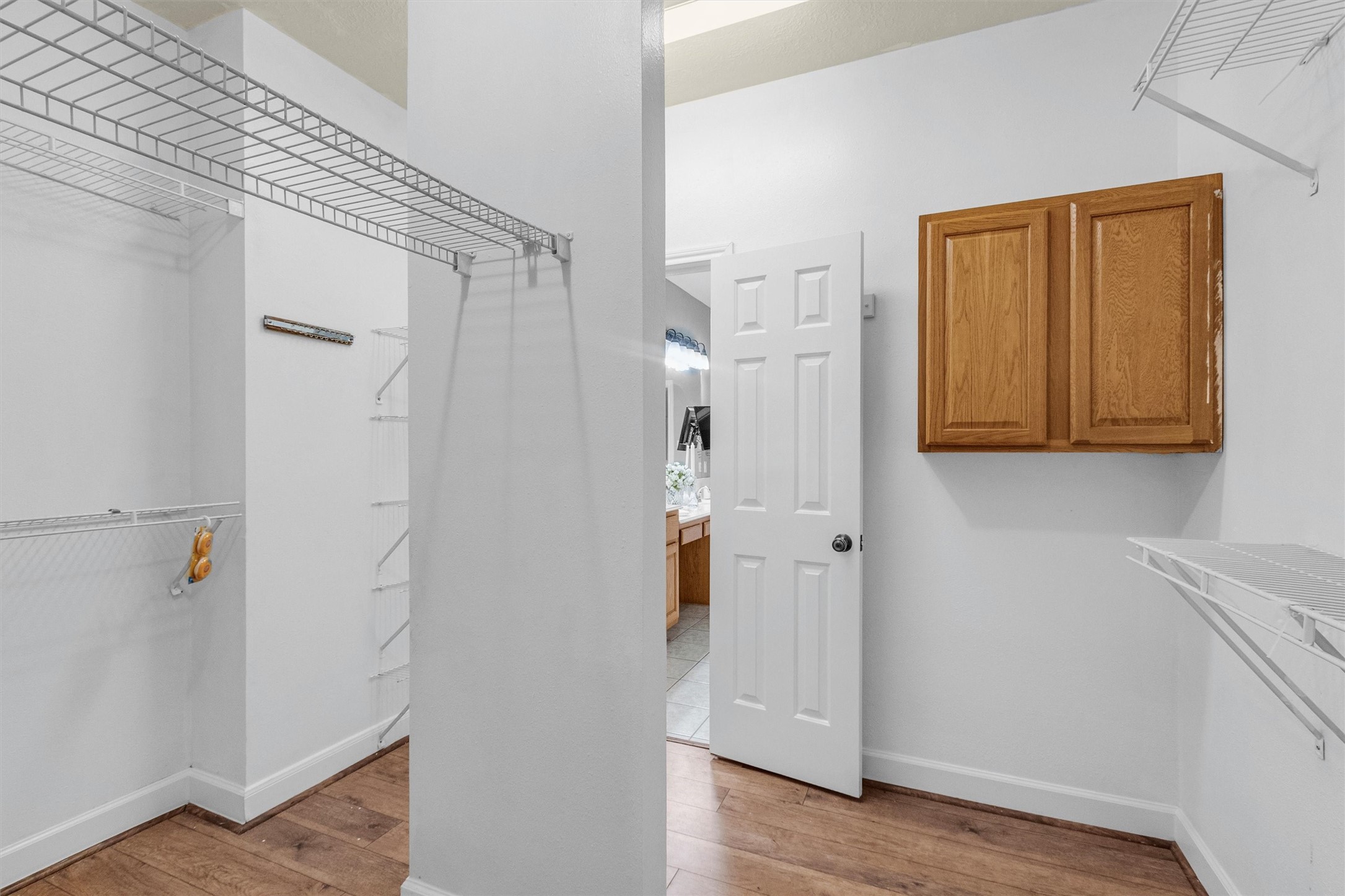 23 Alba Road Montgomery, TX 77356 - Photo 21 of 37 This photo shows a spacious walk-in closet featuring wire shelving and a wooden cabinet for storage. The closet has a door leading to a bathroom with visible vanity and sink. The flooring is wood, and the walls are painted white, providing a clean, open look.