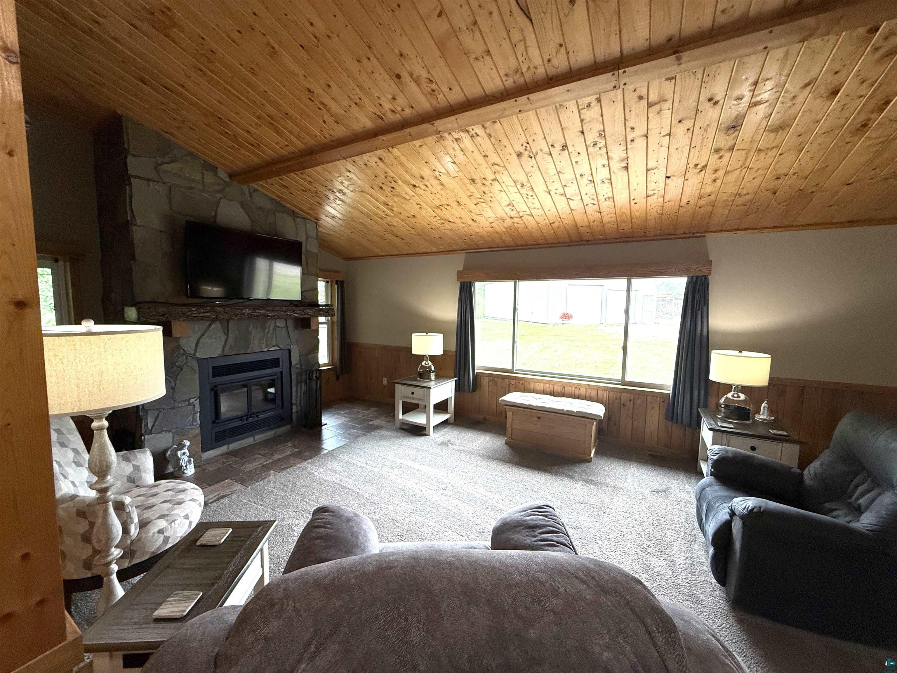 5080 Tower Road Moose Lake, MN 55767 - Photo 9 of 49 Vaulted wood-paneled ceiling, stone-clad fireplace with wood mantle, window with exterior landscape, wood-finish flooring, and wall paneling