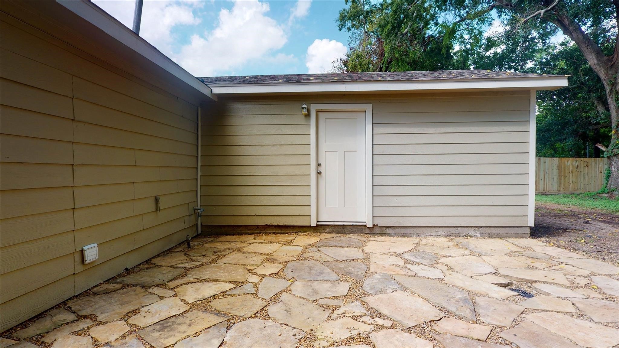 7346 Iwo Jima Road Houston, TX 77033 - Photo 19 of 20 This photo shows a small, simple outdoor area with a light-colored siding exterior and a stone patio. The space is bordered by a wooden fence and features a single door leading into the building. Ideal for low-maintenance outdoor relaxation.