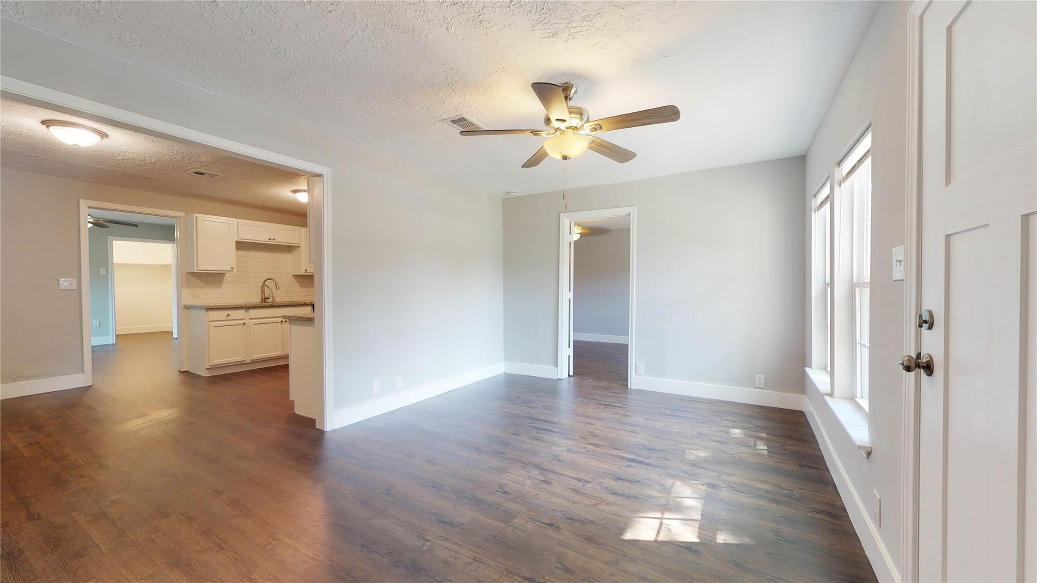7346 Iwo Jima Road Houston, TX 77033 - Photo 2 of 20 This photo showcases a bright, open living area with hardwood floors and a ceiling fan. It connects to a kitchen with white cabinetry and has large windows for natural light.