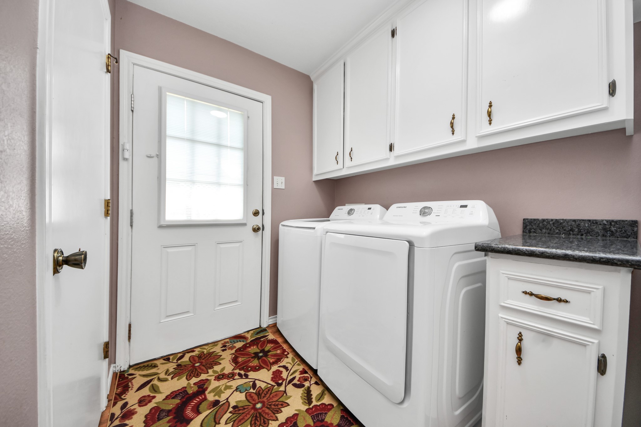 12746 Roy Road Pearland, TX 77581 - Photo 29 of 47 This photo showcases a tidy laundry room featuring a washer and dryer, white cabinetry for storage, and a decorative floral rug. The room has a door with a window, allowing natural light to enter, and a countertop for added convenience.