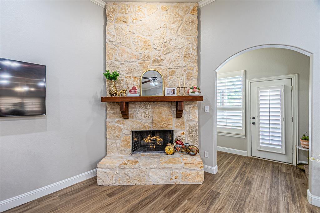 1805 Stetson Way Allen, TX 75002 - Photo 7 of 22 Stone fireplace with a wood mantel, wood-finish flooring, white baseboards, and an arched doorway leading to a door with plantation shutters and a side window with plantation shutters