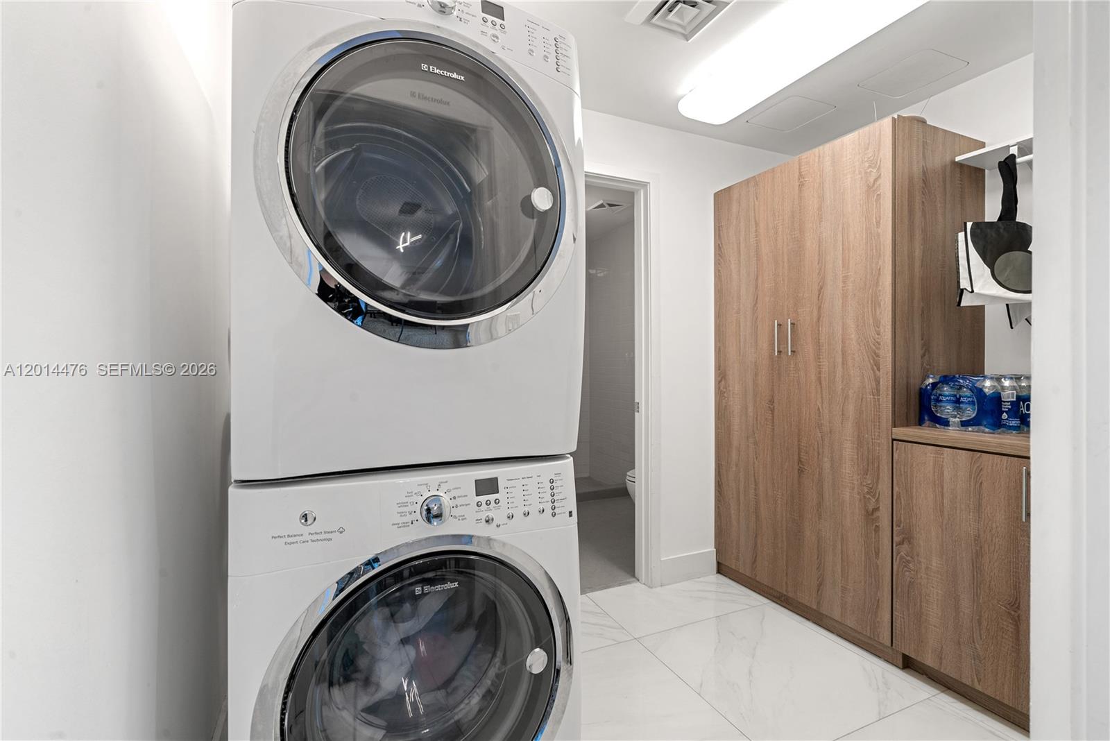 360 Ocean Drive, Unit 1106S Key Biscayne, FL 33149 - Photo 22 of 67 Laundry / Utility Area – A dedicated laundry area features a stacked washer and dryer, glossy marble-look flooring, and built-in wood cabinetry for storage. Bright overhead lighting and a clean layout keep everyday essentials organized and accessible.