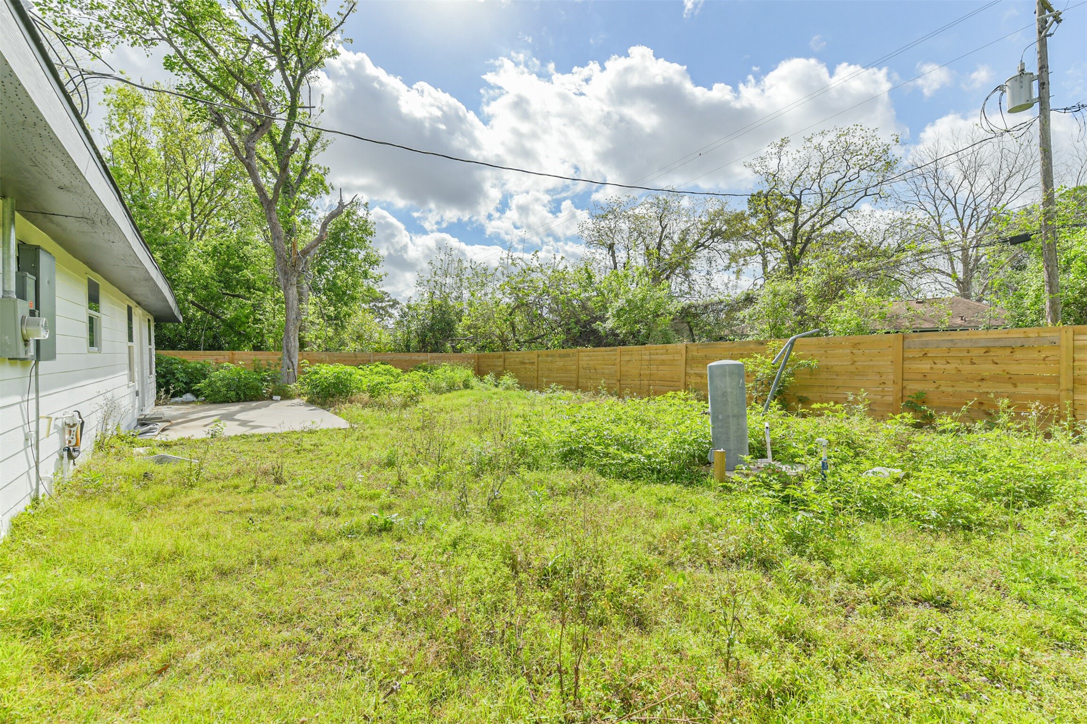 209 Leago Street Houston, TX 77022 - Photo 21 of 24 This photo shows a backyard with a grassy lawn, bordered by a wooden fence. The house has a light exterior with visible utilities. Mature trees provide shade, and there's a small concrete patio area. The setting is spacious and natural, ideal for outdoor activities.