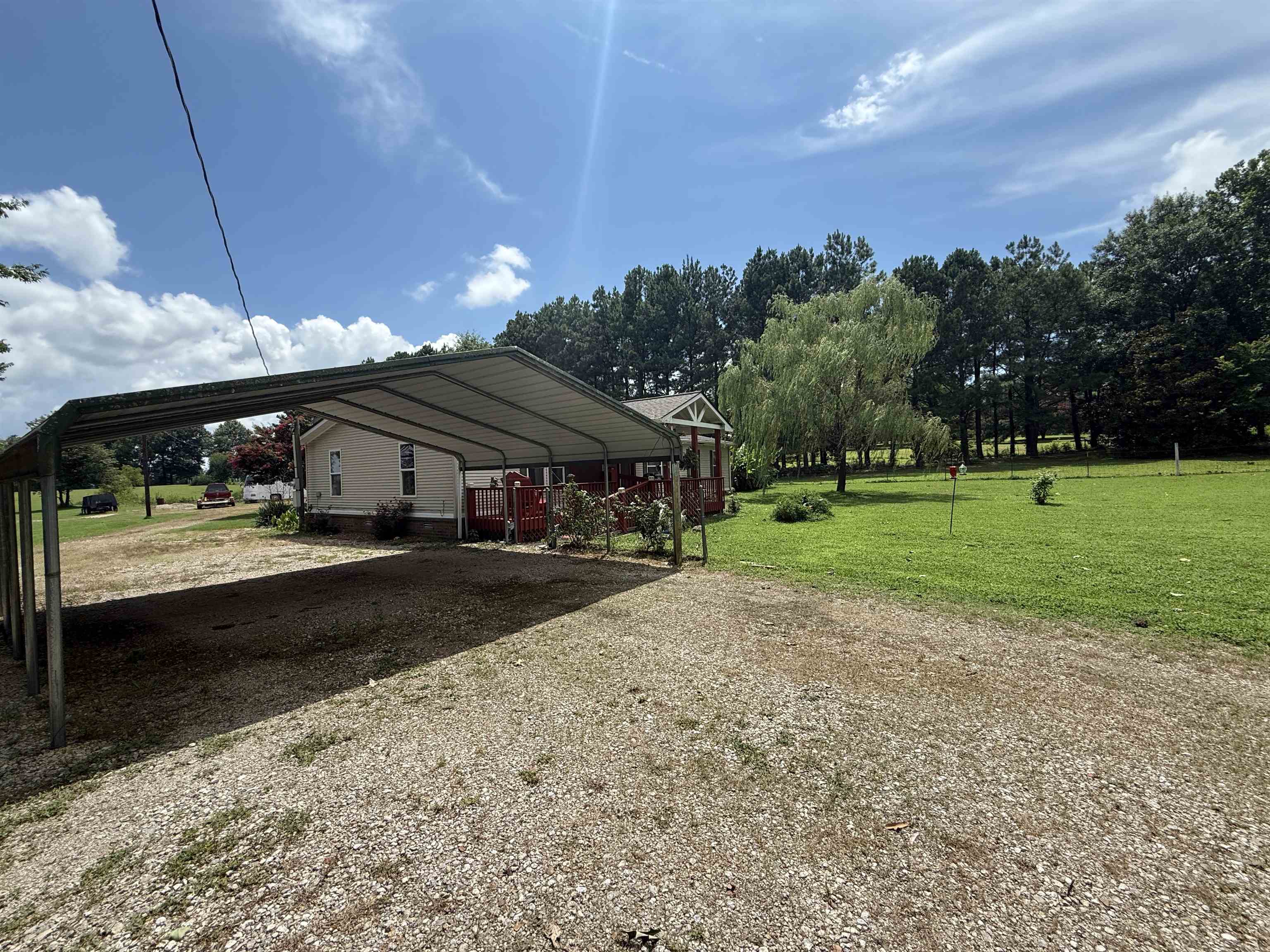 2201 Robert Johnson Road Covington, TN 38019 - Photo 25 of 25 Expansive gravel driveway featuring a metal carport structure, a tan siding residence with a red deck, a manicured green lawn, and mature trees bordering the property