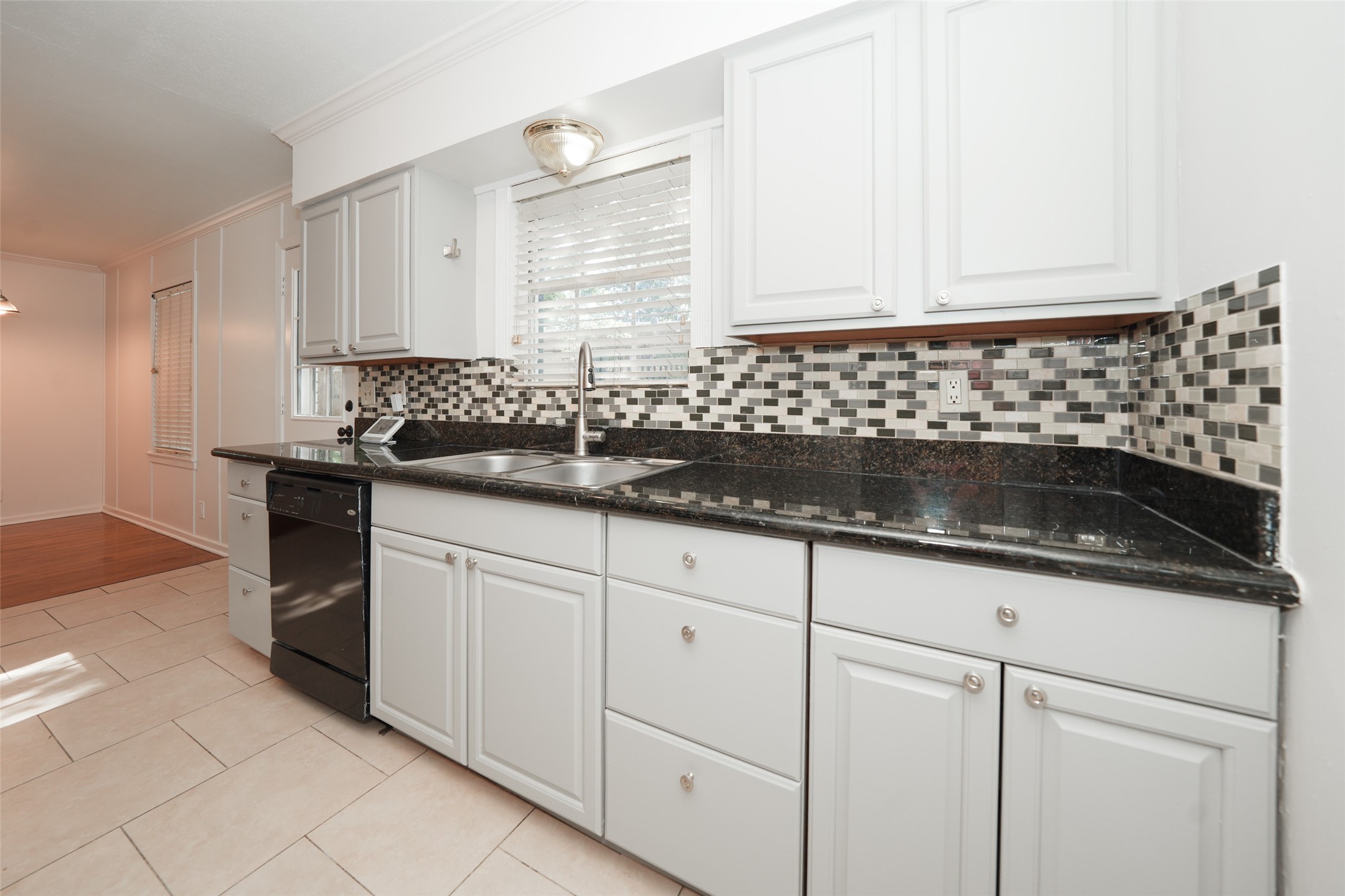 9519 Val Verde Street Houston, TX 77063 - Photo 13 of 27 This kitchen features modern white cabinetry, sleek black countertops, and a stylish mosaic tile backsplash. It includes a built-in dishwasher and a double sink beneath a window, allowing for plenty of natural light. The space seamlessly connects to the dining area with a combination of tile and wood flooring.