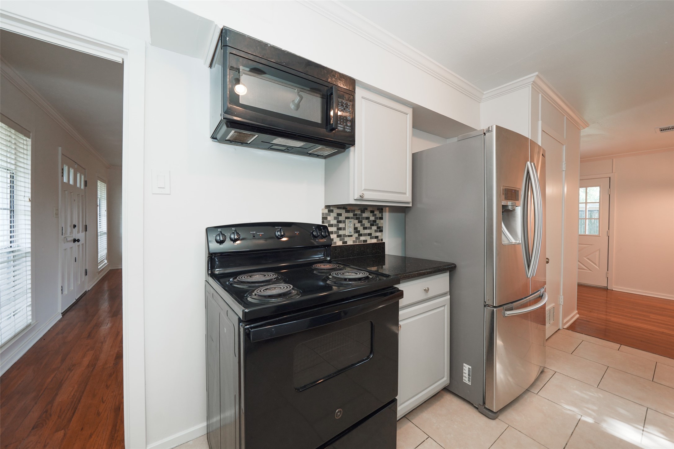9519 Val Verde Street Houston, TX 77063 - Photo 14 of 27 This kitchen features a black stove and microwave, stainless steel refrigerator, and modern tile backsplash. It opens into a hallway with wood flooring and ample natural light from nearby windows.