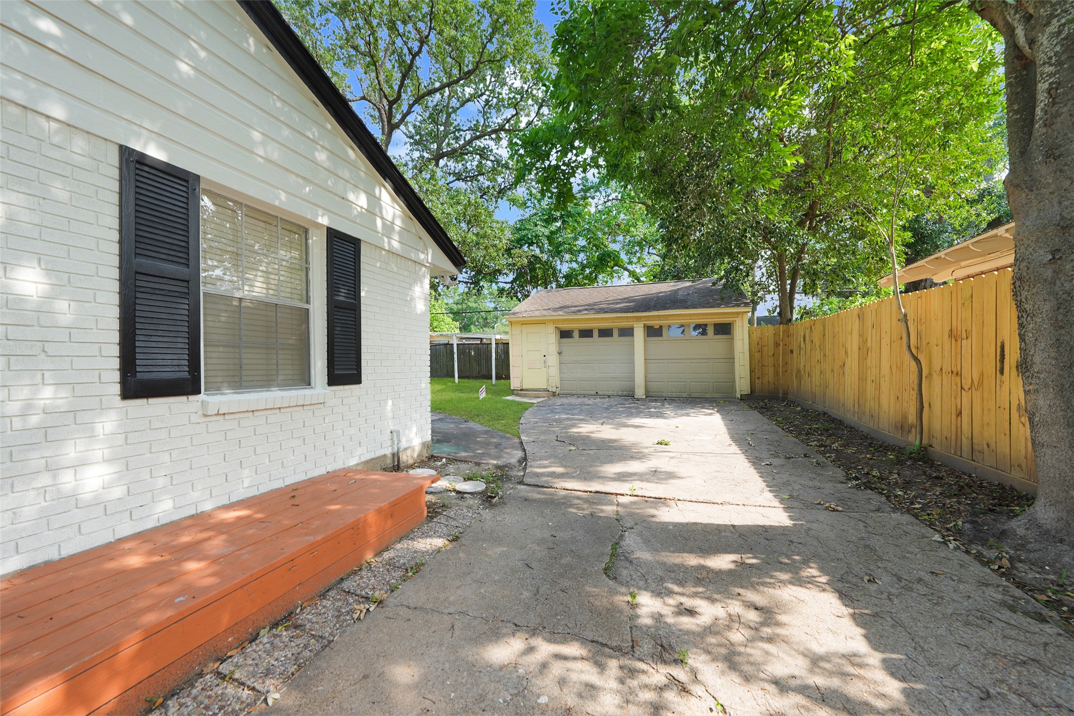 9519 Val Verde Street Houston, TX 77063 - Photo 3 of 27 This photo shows a charming backyard view featuring a white brick house with black shutters, a wooden step, and a spacious driveway leading to a detached two-car garage. The area is partially shaded by mature trees and bordered by a wooden fence, offering a blend of privacy and natural beauty.