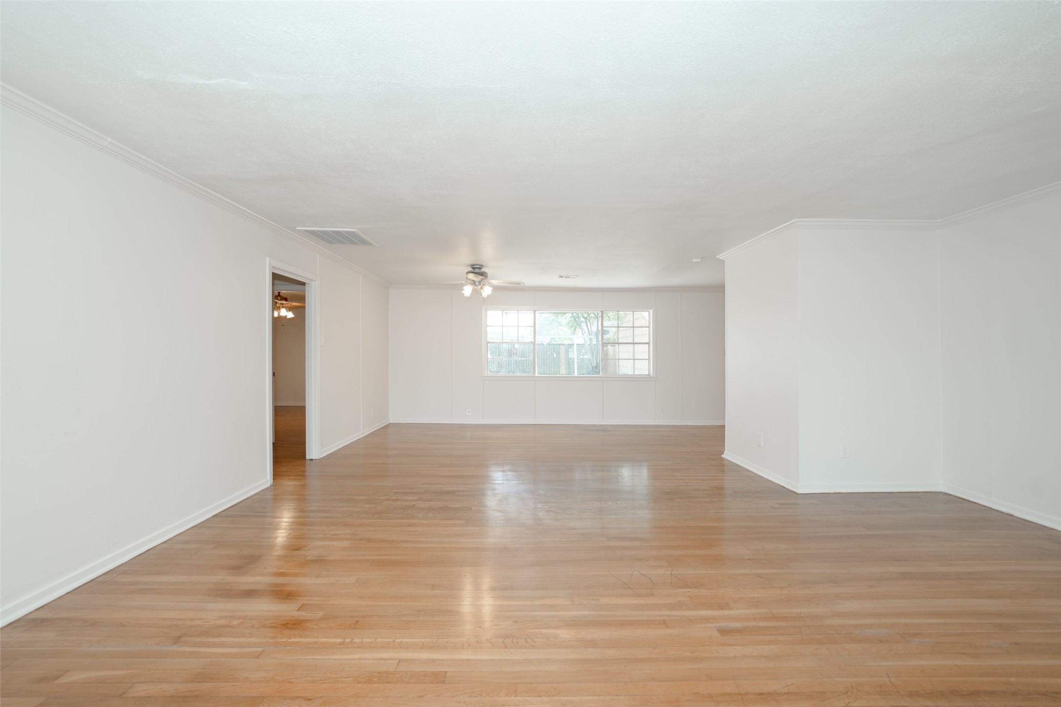 9519 Val Verde Street Houston, TX 77063 - Photo 10 of 27 This photo showcases a spacious, bright living area with light hardwood floors and white walls. A large window allows natural light to fill the room, and a ceiling fan is centrally located. An open doorway leads to an adjacent room, offering a versatile layout for a home buyer.