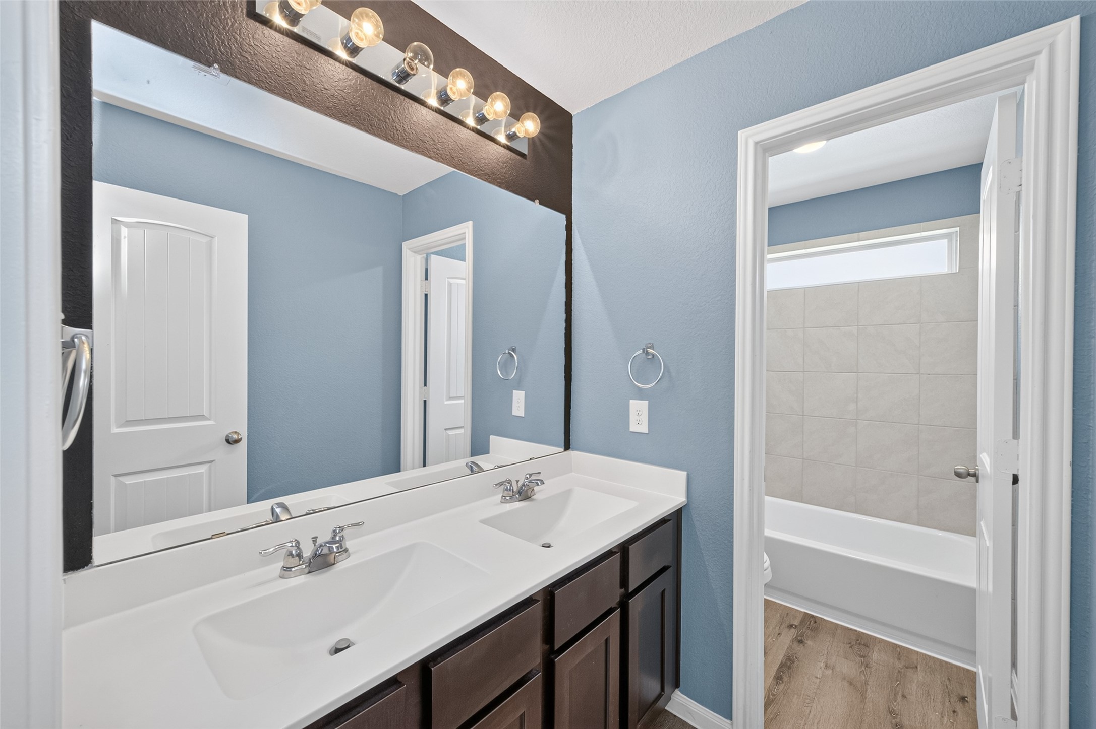 2526 Sturgeon Way Conroe, TX 77384 - Photo 33 of 49 This bathroom features a double vanity with dark wood cabinets, a large mirror, and modern lighting. The walls are painted a soothing blue, and there's an adjacent room with a tiled bathtub. The space is well-lit and has a clean, contemporary design.