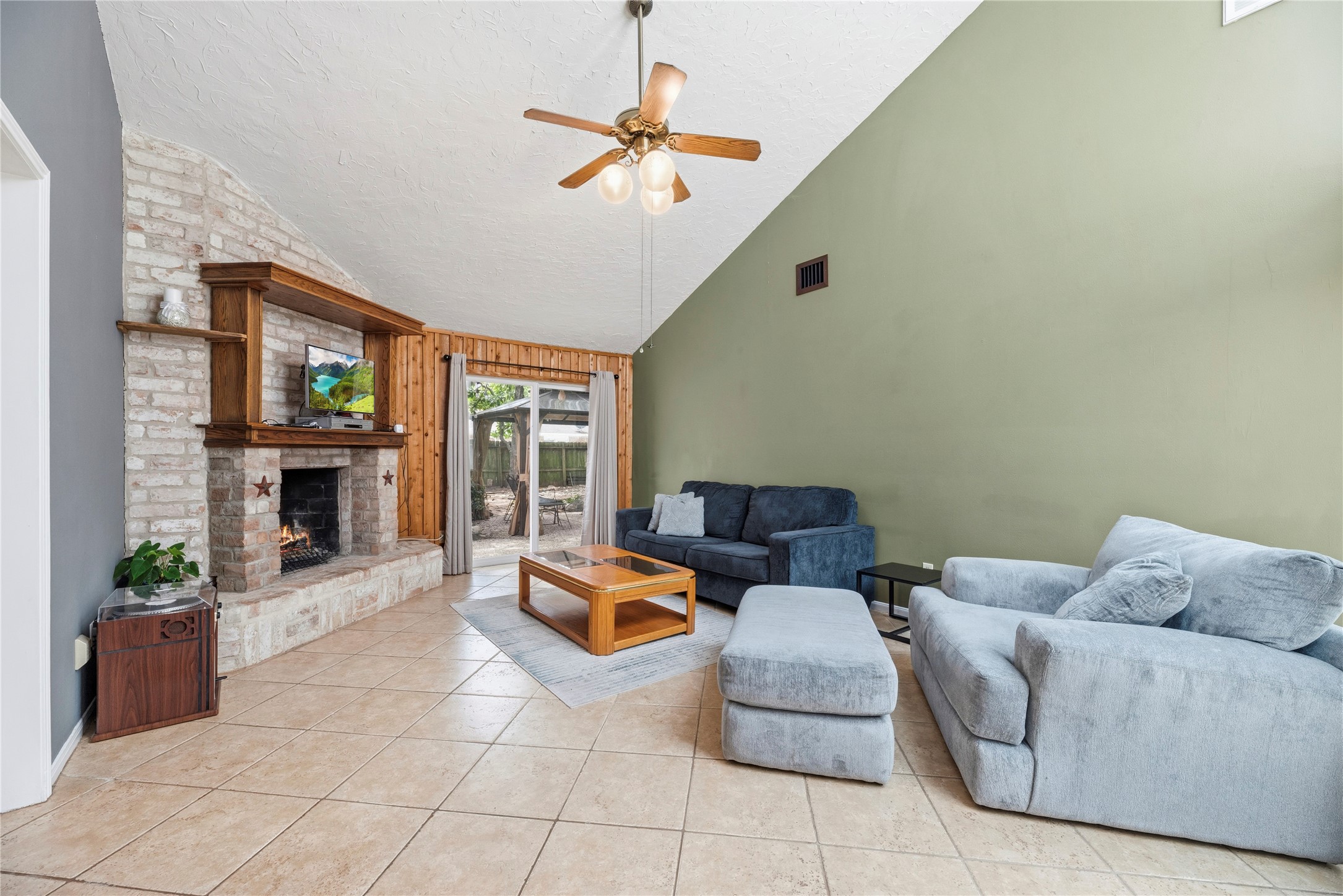 14203 Wickersham Lane Houston, TX 77077 - Photo 9 of 25 Grand living room featuring high vaulted ceilings, a ceiling fan, and a floor-to-ceiling white brick fireplace with a wood mantel. Sliding glass doors open directly to the back patio.