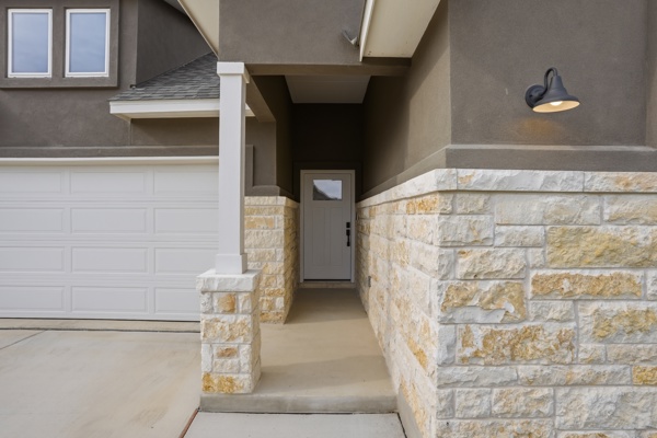 201 Prodigal Way Kyle, TX 78640 - Photo 4 of 22 The property features a stone and stucco exterior, a white panel garage door, and a covered entryway with an exterior light fixture