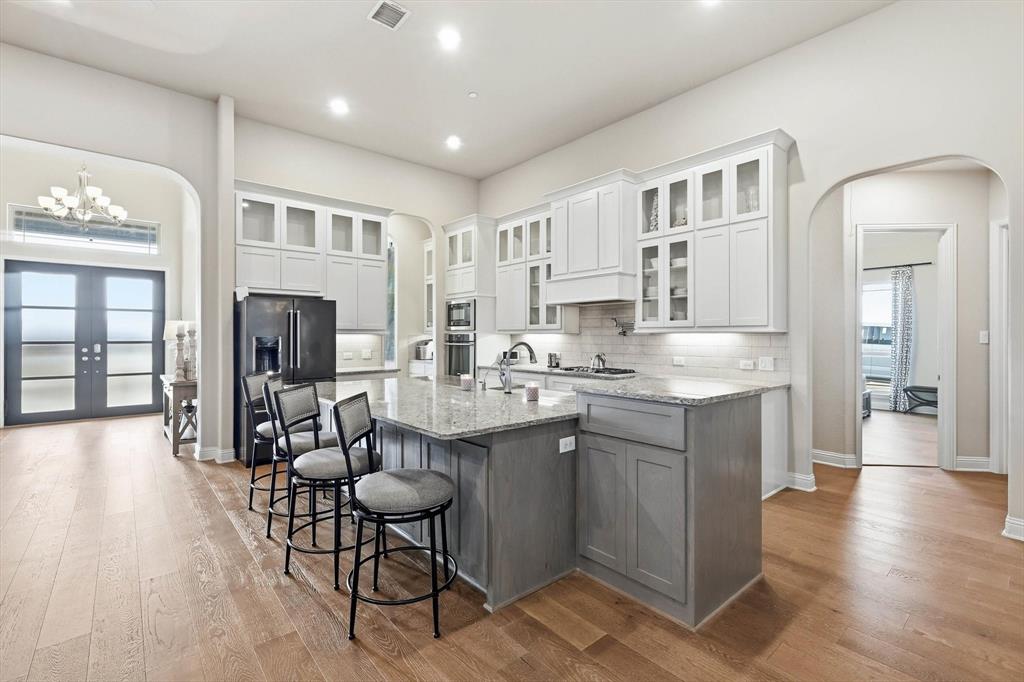 7751 Faught Road Northlake, TX 76226 - Photo 12 of 39 Open-concept kitchen featuring an oversized island with seating, light wood-finish flooring, white upper cabinetry with glass fronts, and a subway tile backsplash